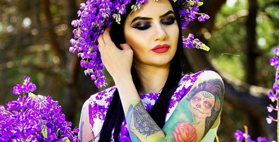 mulher com cabelo escuro segurando flores roxas e com uma coroa de flores roxas que tem uma tatuagem de mulher esquelética La Cartina no braço, uma tatuagem tradicional mexicana para mulheres