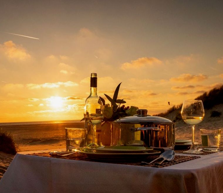 mesa de jantar na praia com prato e taça de vinho, sol se pondo ao fundo